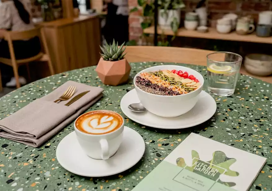 Smoothie bowl topped with pumpkin seeds, pomegranate seeds, and grains served beside a latte with latte art.
