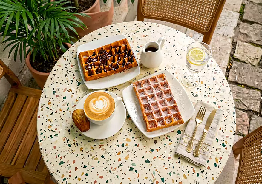 Two waffles, one drizzled with dark sauce and one dusted with powdered sugar, served with a cup of coffee and a small milk pitcher.