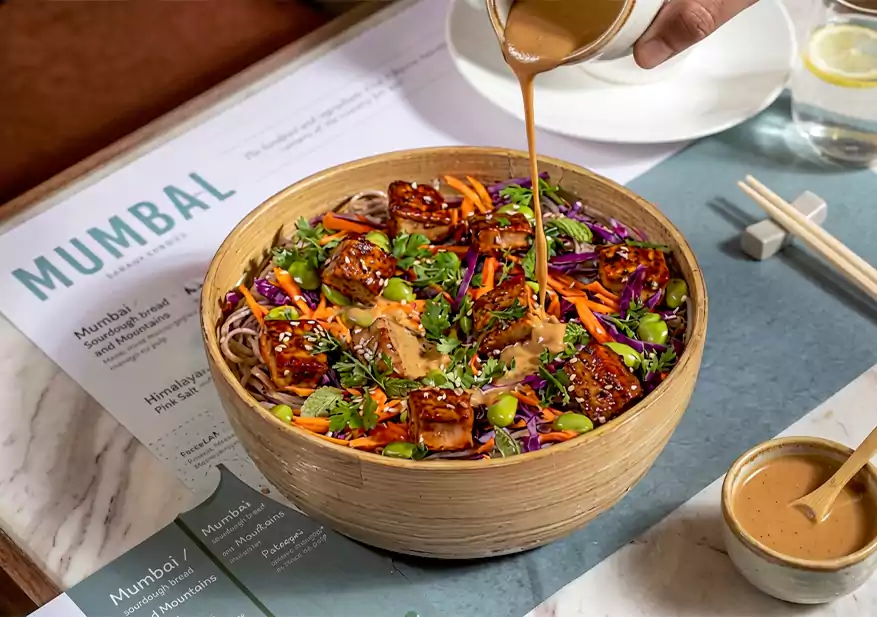 Wooden bowl with noodles, shredded carrots, purple cabbage, tofu cubes, and sauce being poured over the dish.